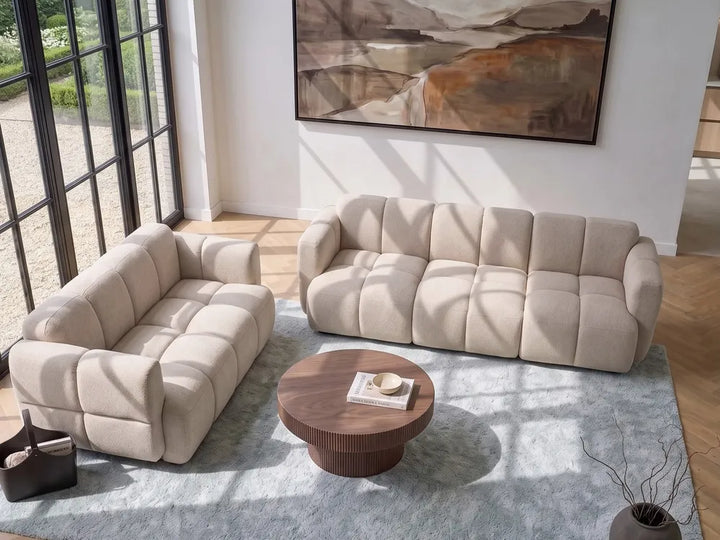 A modern living room setup featuring a beige tufted loveseat and a matching beige three-seater sofa arranged on a light blue-gray rug around a round wooden coffee table. Large black-framed windows on the left let in natural light, and an abstract painting hangs on the white wall above.