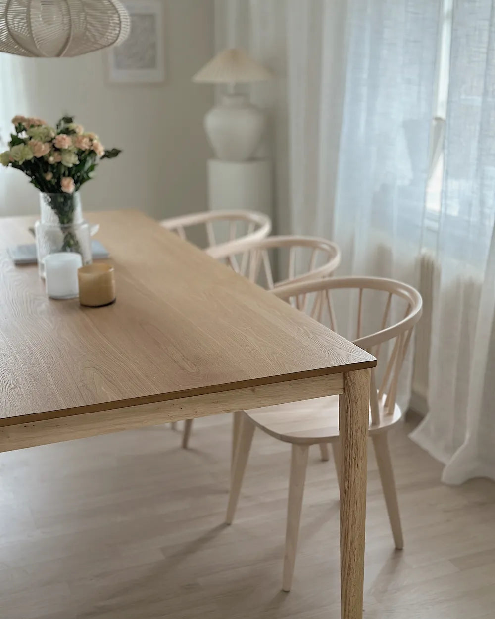 Dining room with wooden table and chairs, flowers, and a lamp in a bright room.