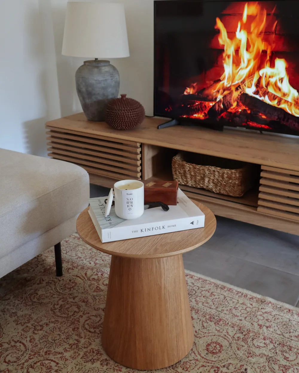 Living room with a wooden side table, lamp, and TV displaying a fire.