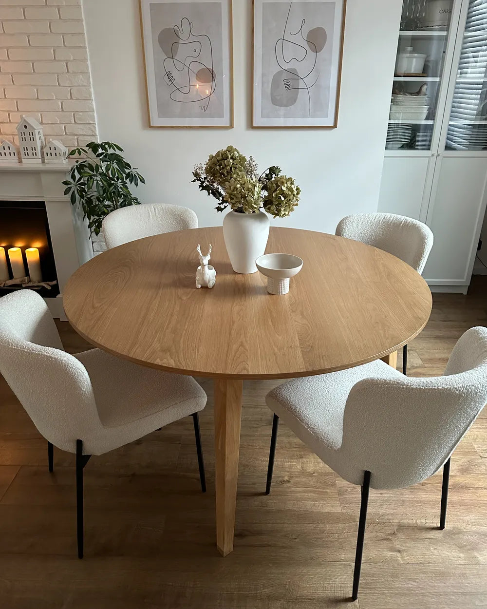 Round wooden dining table with four white chairs in a room with framed art on the wall.