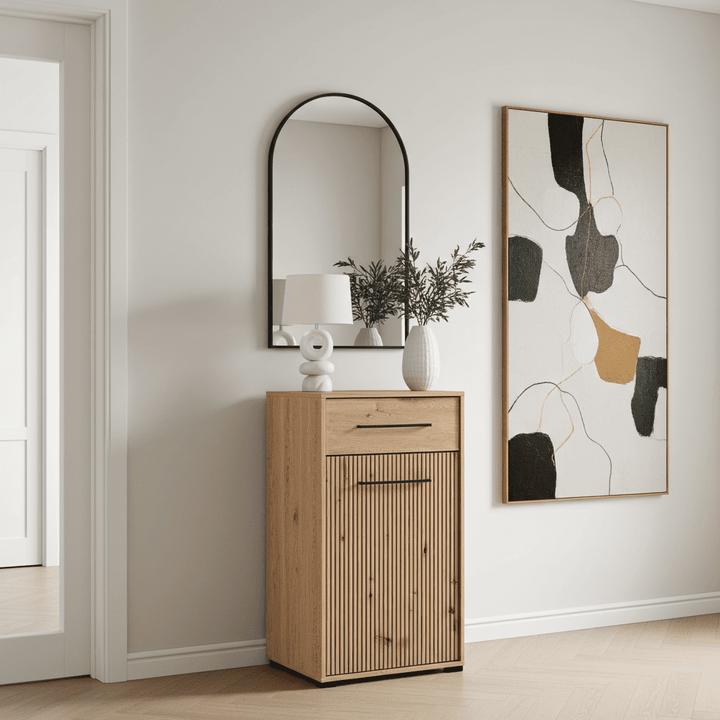 A light natural wood entryway cabinet with a ribbed door and a single drawer, styled in a minimalist room with a black-framed arched mirror, a white table lamp, and a vase. To the right is a large abstract framed wall art piece on a white wall.
