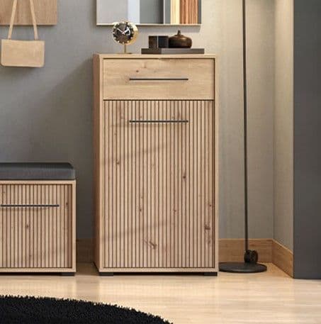 A tall light wood entryway cabinet with a slatted front door and a top drawer, shown in a styled room setting next to a bench and floor lamp.