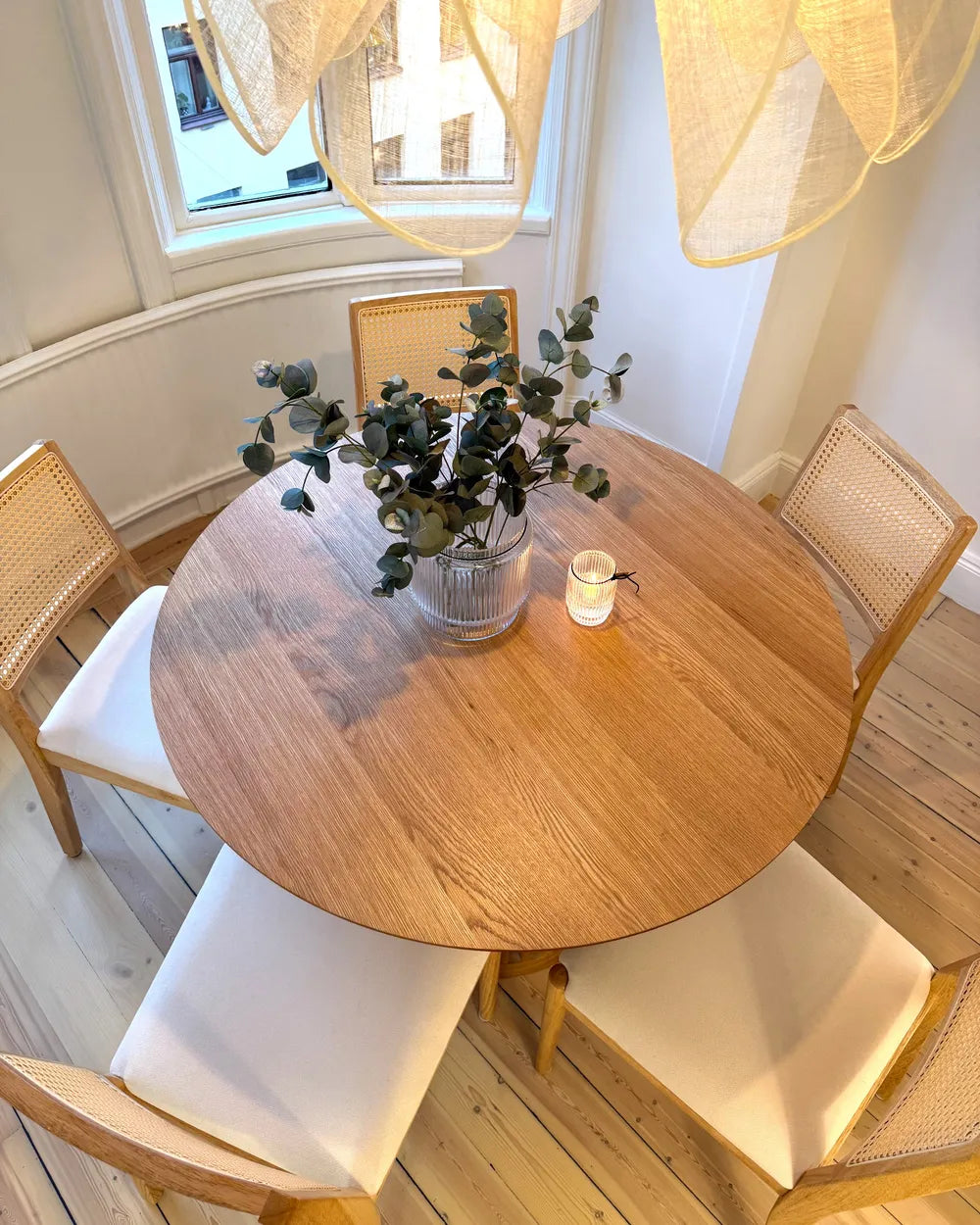 Round wooden dining table with chairs and a plant in a room with large windows.