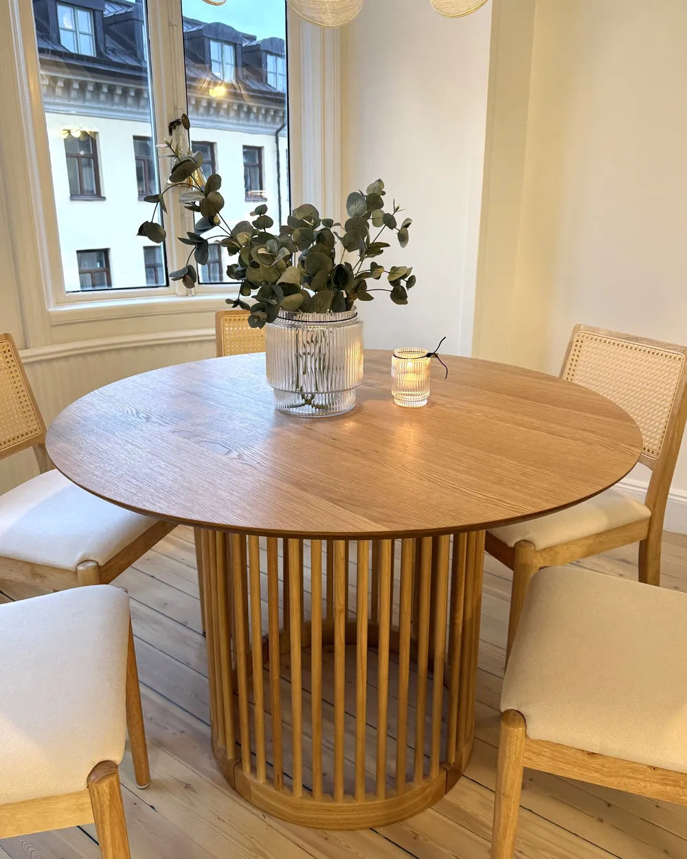 Round wooden dining table with chairs and a plant in a room with large windows.