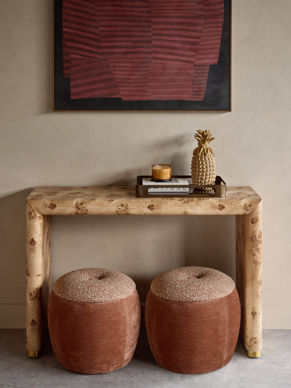 A rustic console table made from natural burl wood, styled with two round terracotta poufs, a small tray, a candle, and a decorative pineapple on top, set against a neutral beige wall with framed abstract art.