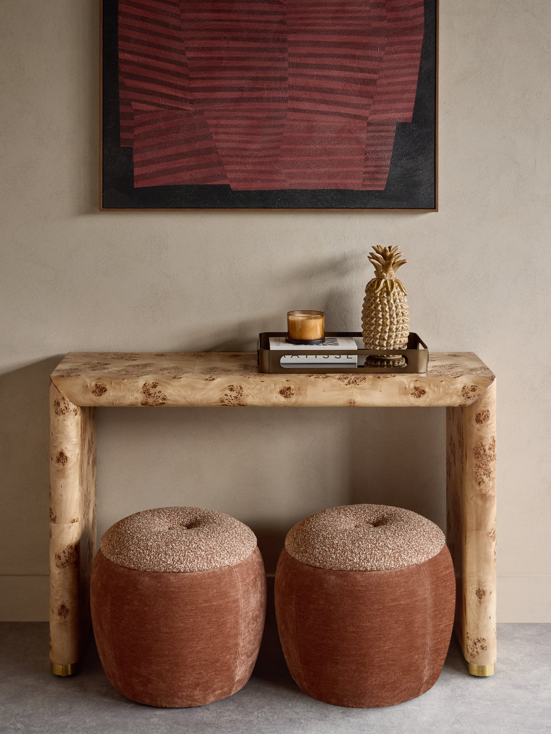 A rustic console table made from natural burl wood, styled with two round terracotta poufs, a small tray, a candle, and a decorative pineapple on top, set against a neutral beige wall with framed abstract art.