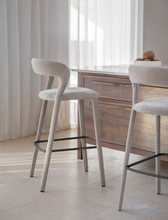 A modern bar stool with a curved backrest, upholstered in light beige fabric, set in a bright, minimalist kitchen or dining area next to a wooden island.