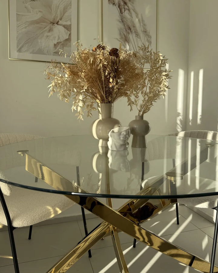Glass dining table with gold legs, chairs, and decorative elements in a room with light walls.