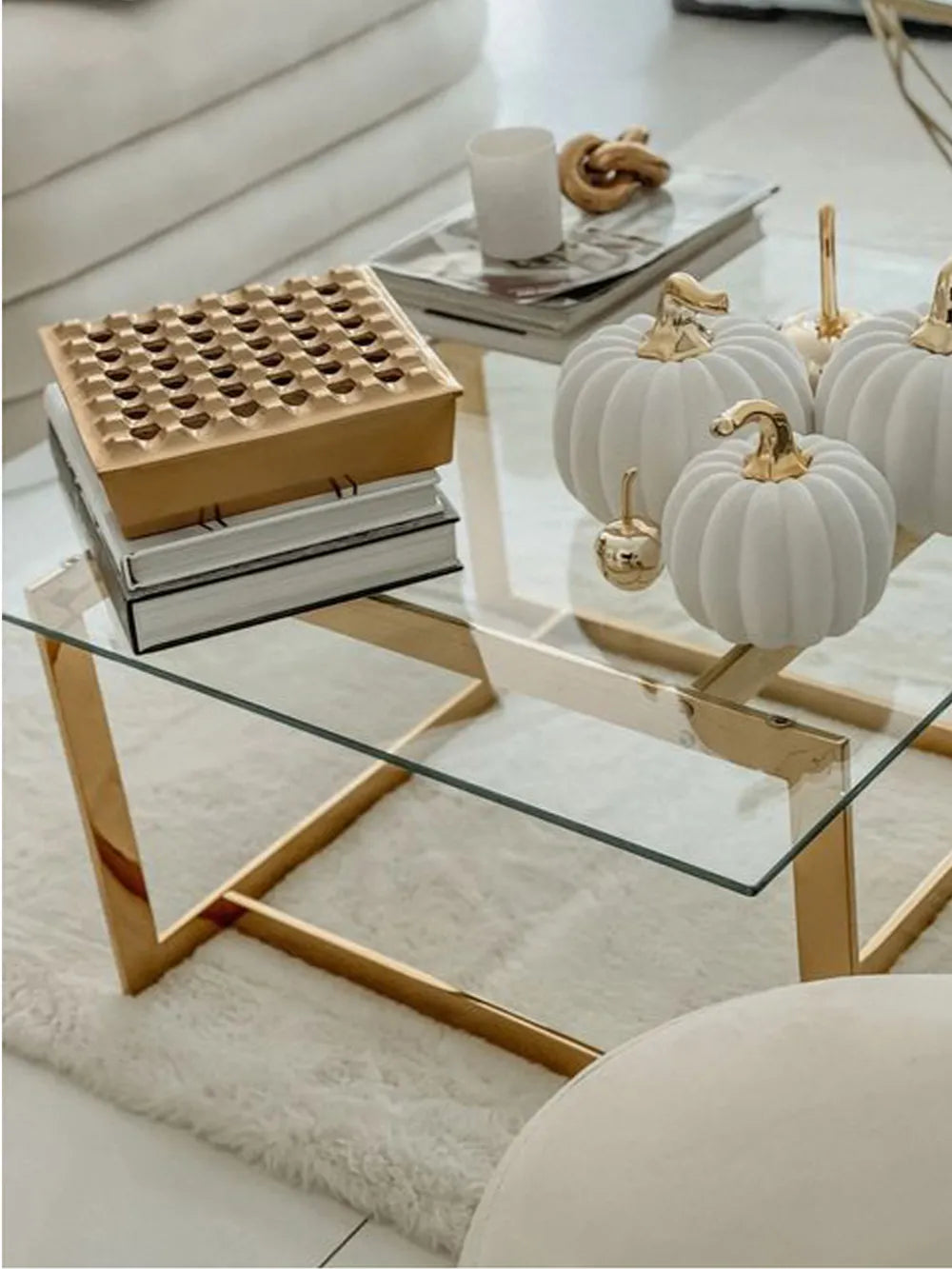 Glass coffee table with decorative items including a wooden puzzle, books, and white pumpkins with gold accents.