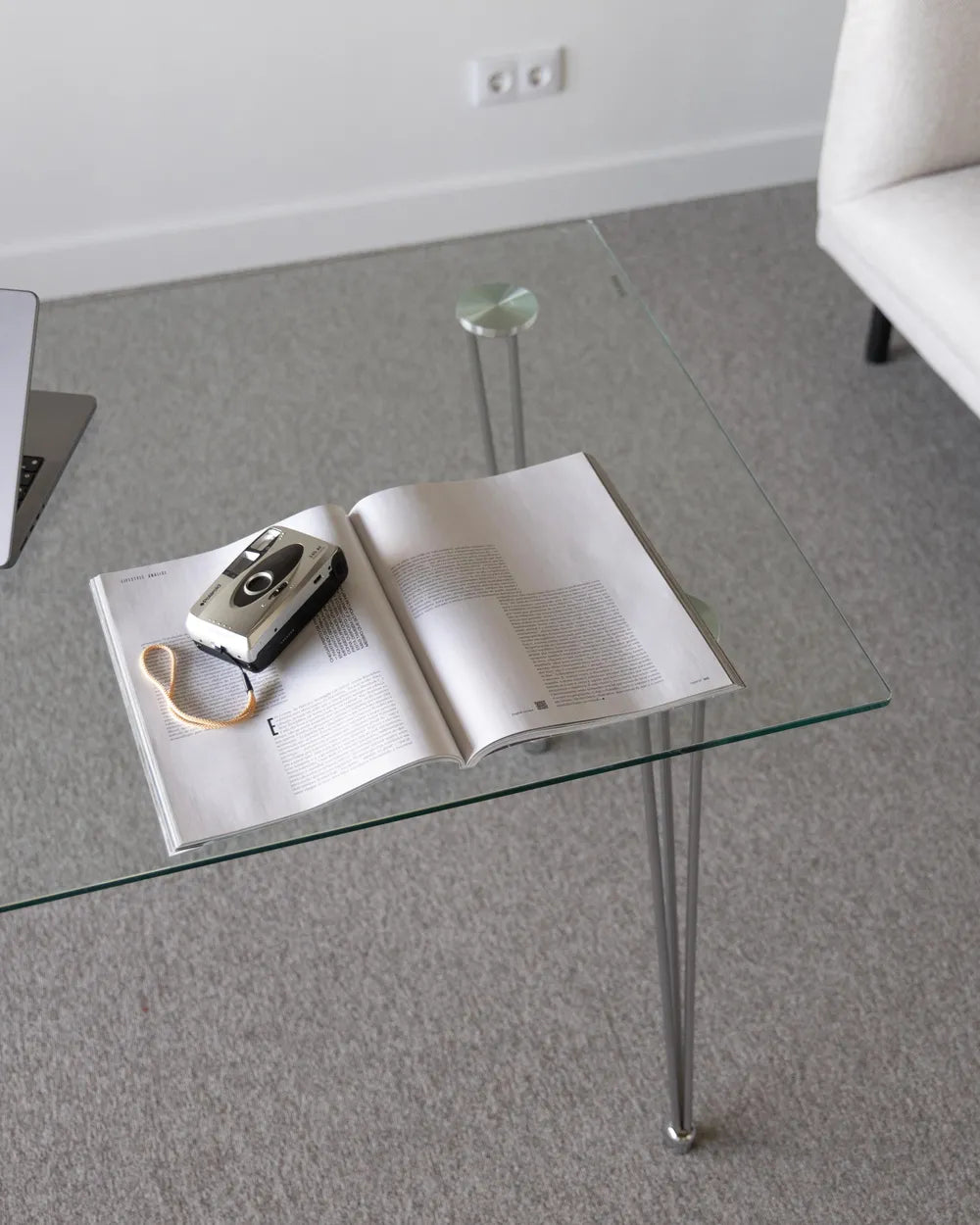 Glass coffee table with an open book and camera on a gray floor.