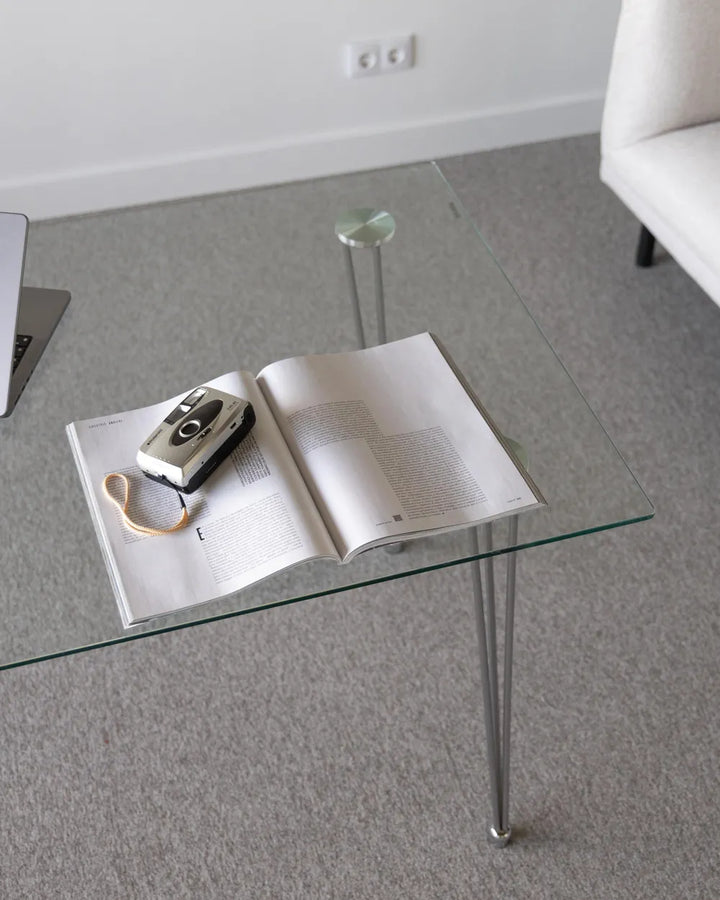 Glass coffee table with an open book and camera on a gray floor.