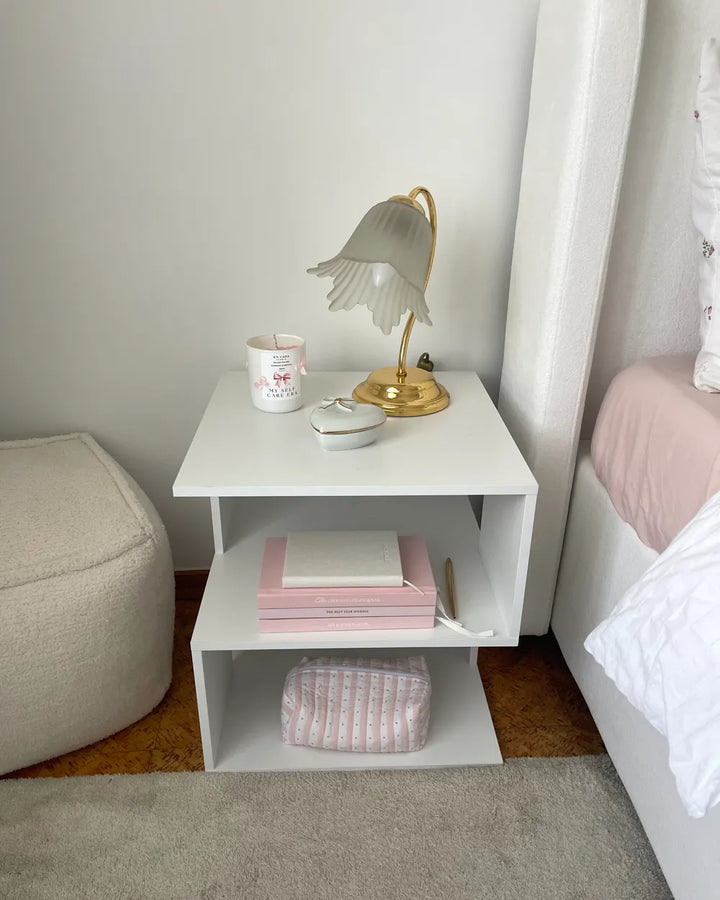 A modern white two-tier nightstand styled in a bedroom corner, holding a gold-framed table lamp, a white mug, and a small white bowl. The nightstand is positioned between a bed with pink bedding and a beige ottoman.