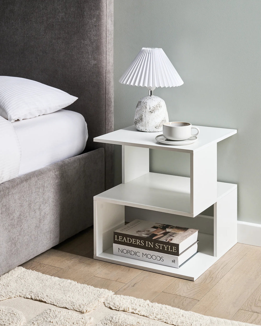 White nightstand with lamp and books next to a bed in a bedroom.