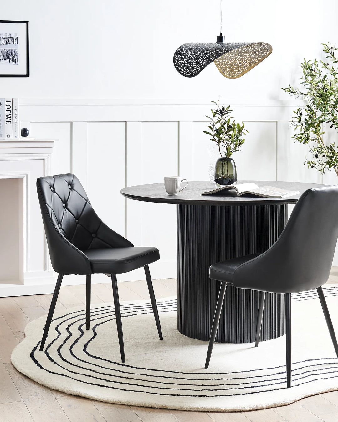 A modern dining room scene featuring a round black ribbed table, two black upholstered chairs, a white patterned rug, and a black pendant light in a bright, white-walled room.