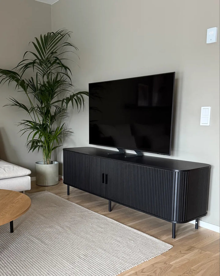 A modern black pleated TV stand cabinet with legs, holding a large black flat-screen TV, set in a living room with a beige wall, wood floor, a potted palm, and a textured rug.