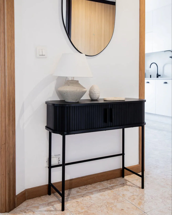A modern black console table with a ribbed front and slim metal legs, styled in a home entryway with a white wall, wood trim, a mirror, and a table lamp.