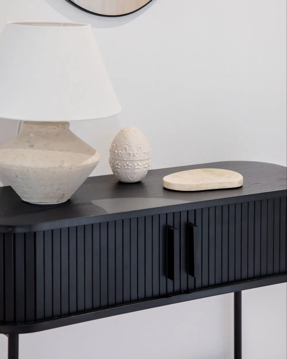 A modern black console table with vertical fluted detailing and black handles, styled with a textured white table lamp, a small carved stone egg, and a round wooden tray against a light gray wall.