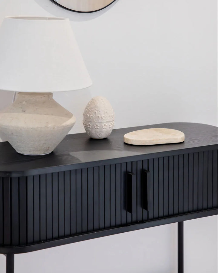 A modern black console table with vertical fluted detailing and black handles, styled with a textured white table lamp, a small carved stone egg, and a round wooden tray against a light gray wall.