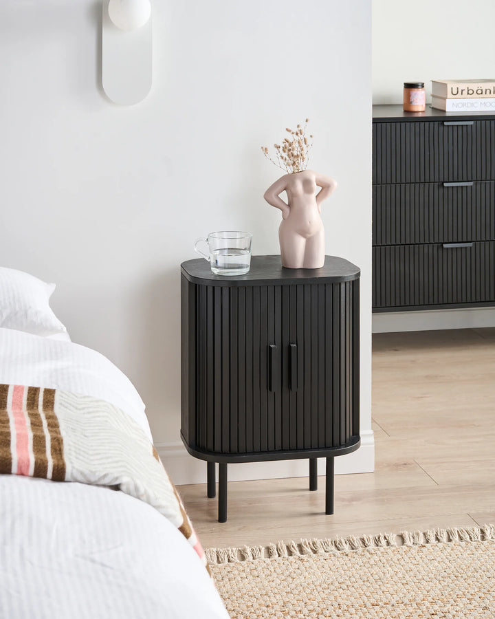 A modern black nightstand with vertical fluted detailing and tapered legs, styled in a bedroom next to a bed with white linens. On top of the nightstand sits a small pink ceramic figurine holding dried stems and a clear glass cup.