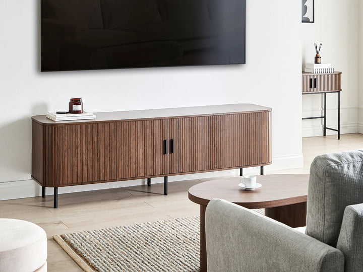 A mid-century modern wooden TV stand in a warm brown finish with vertical slats, black legs, and black handles, styled in a living room setting under a wall-mounted television.
