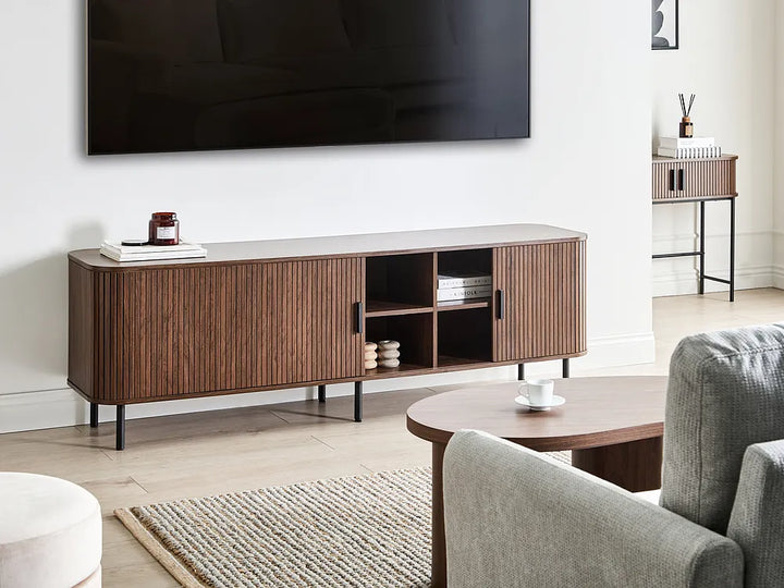 A modern wooden TV console with fluted doors and open shelving, styled in a contemporary living room setting with a wall-mounted TV, armchair, and coffee table.
