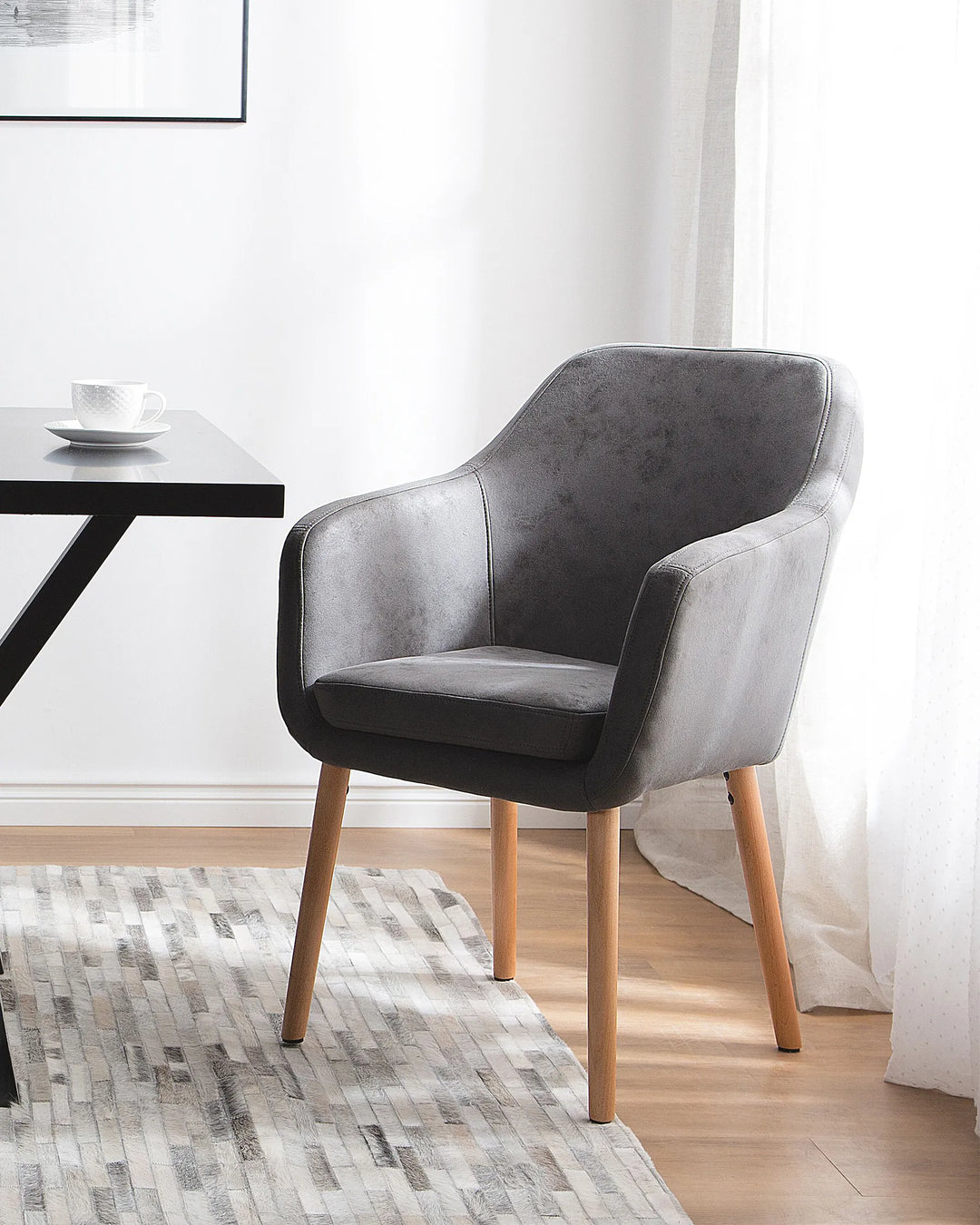 A modern gray upholstered accent chair with light wood legs, styled in a bright room next to a black table on a patterned rug.