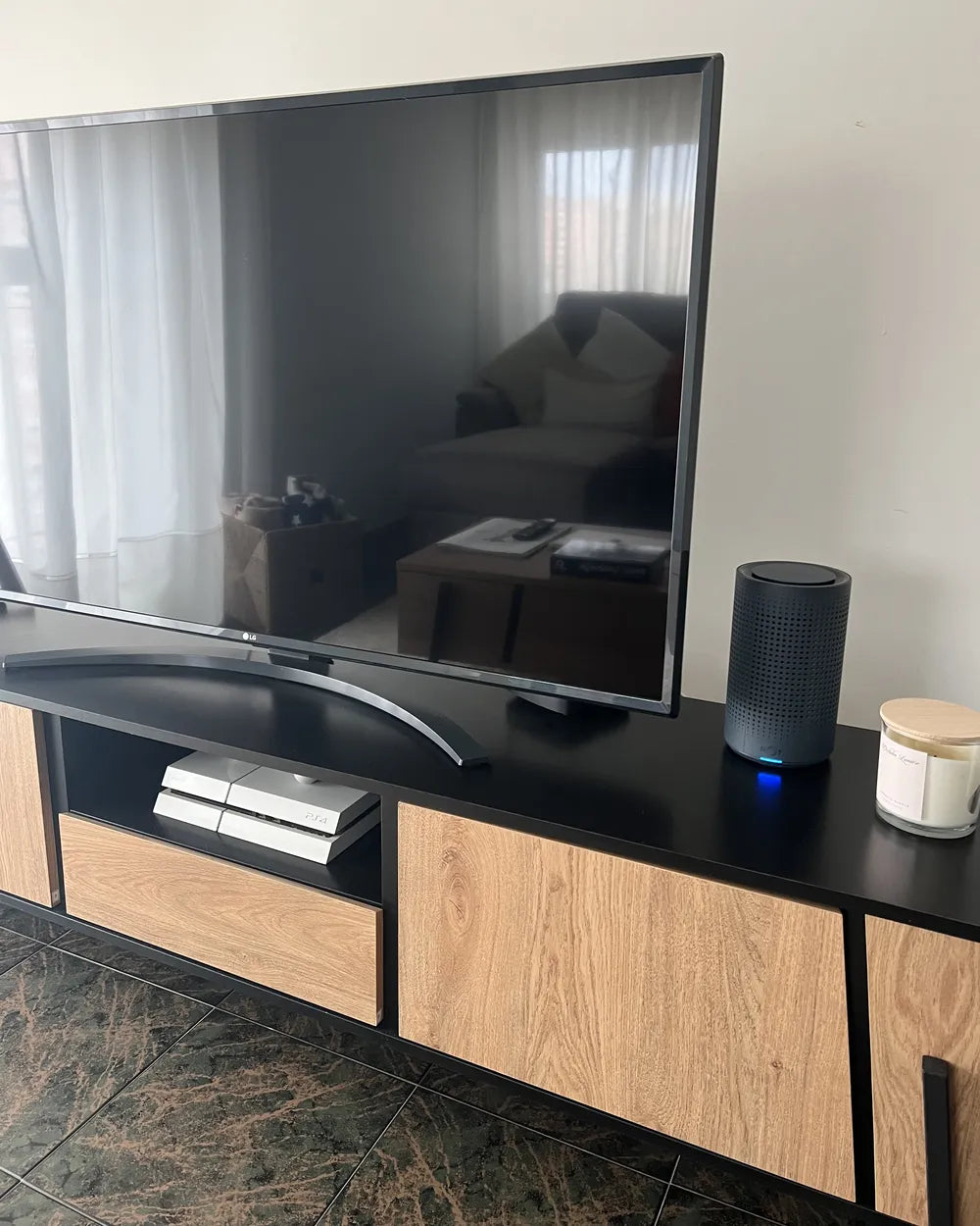 A modern black TV stand with light wood grain cabinets, holding a large flat-screen TV, a black cylindrical smart speaker, and a white candle, set against a neutral indoor wall.