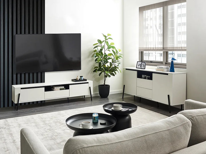 A modern living room setup featuring a white and black media console with a large black TV, a matching white sideboard with a plant, and a light beige sofa in the foreground.