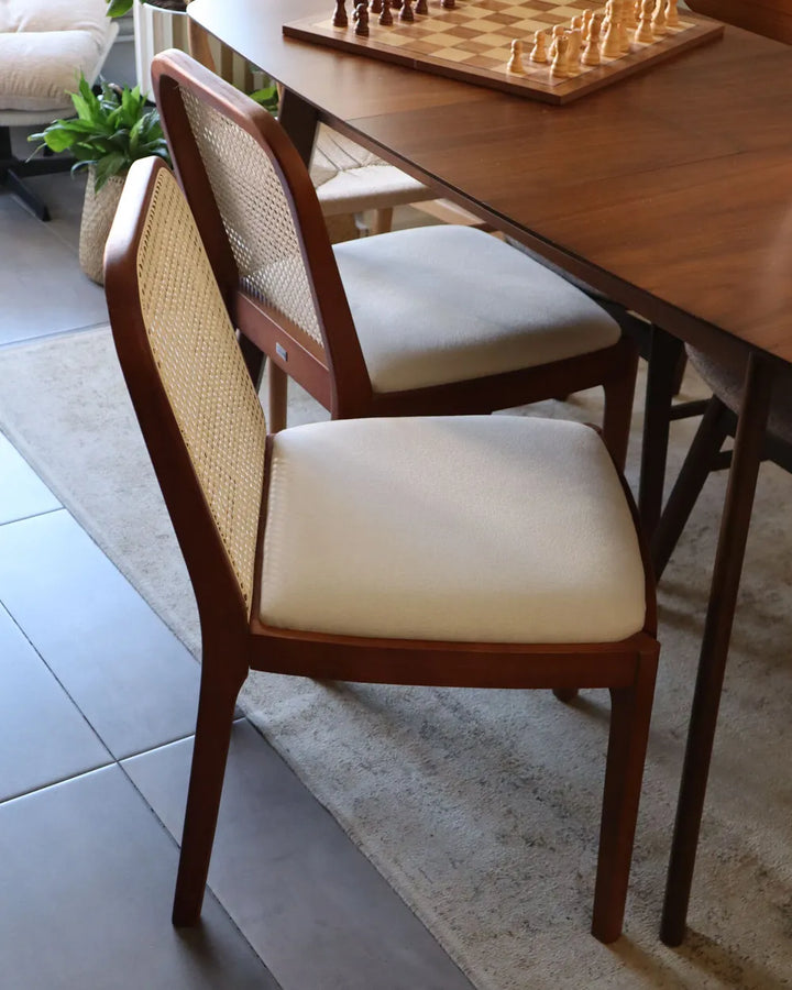 A mid-century modern dining chair with a warm brown wooden frame and woven cane backrest, paired with a cream upholstered seat cushion, shown in a dining room setting next to a wooden table.