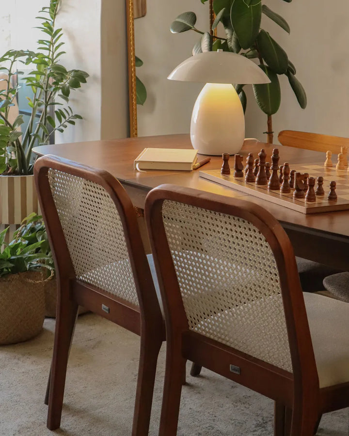 A warm, modern dining room scene featuring two wooden dining chairs with woven cane backs, set around a matching wooden table with a white table lamp, a chessboard, and potted plants.