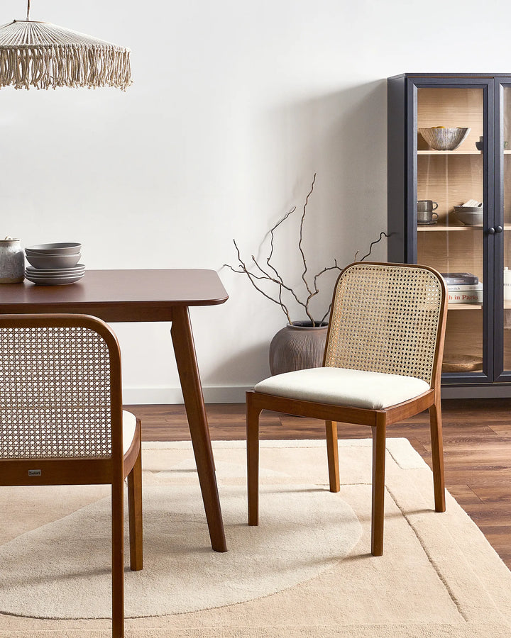 A mid-century modern dining room scene featuring a wooden dining table and two matching chairs with woven cane backs and cream-colored seat cushions, set on a light beige rug against a white wall.