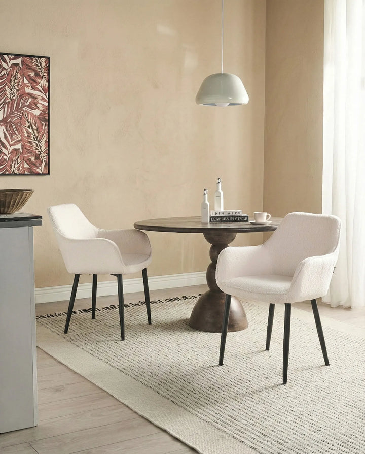 A dining area scene with two upholstered armchairs around a round pedestal table on a woven rug, set in a warm neutral room with a hanging pendant light and wall art.