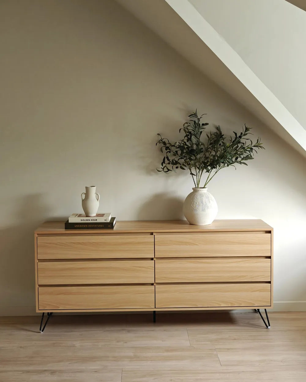 A light natural wood dresser with six drawers and black metal legs, styled in a minimalist room with a slanted ceiling, a white textured vase with greenery, and a small white ceramic pitcher on top.