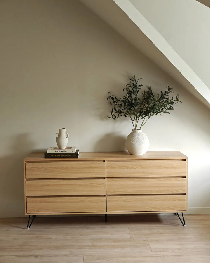 A light natural wood dresser with six drawers and black metal legs, styled in a minimalist room with a slanted ceiling, a white textured vase with greenery, and a small white ceramic pitcher on top.