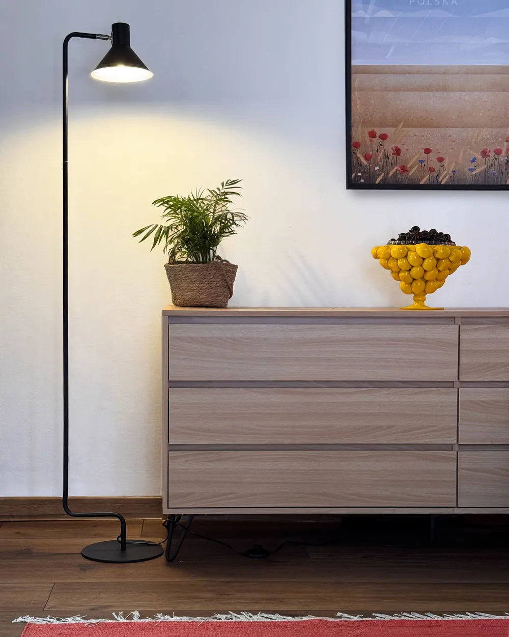 A modern black floor lamp stands next to a light wood dresser in a styled room setting with a potted plant and a yellow bowl of lemons, against a white wall.