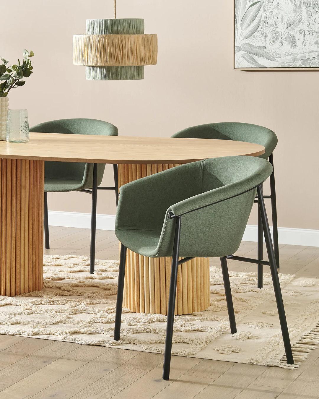 A dining room scene with a light wood oval table and three upholstered chairs in muted sage green, set on a cream rug against a beige wall. A hanging woven pendant light and framed wall art are visible.