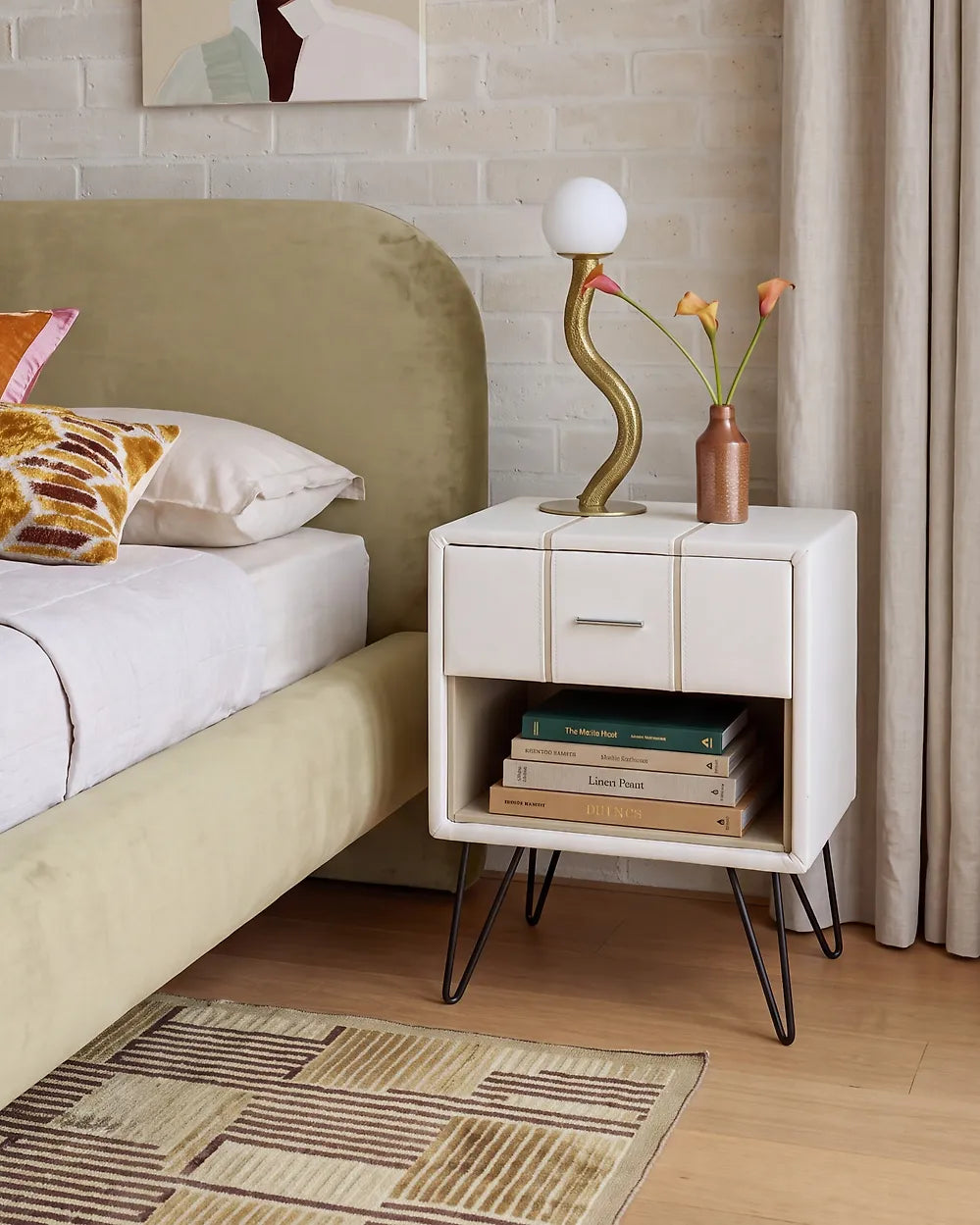 A modern white nightstand with black hairpin legs in a bedroom setting, styled next to a bed with a green headboard, holding books and a decorative lamp.