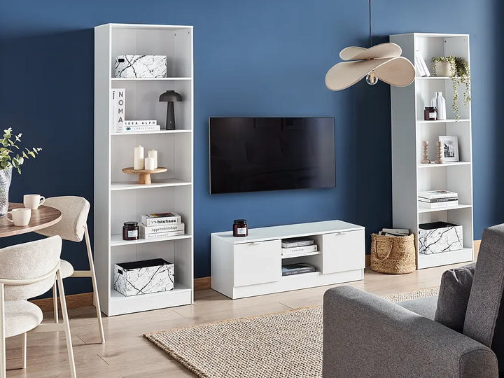 A modern living room setup featuring a white media console, a black flat-screen TV mounted on a blue wall, and two matching tall white bookshelves decorated with books, boxes, and small decor items.