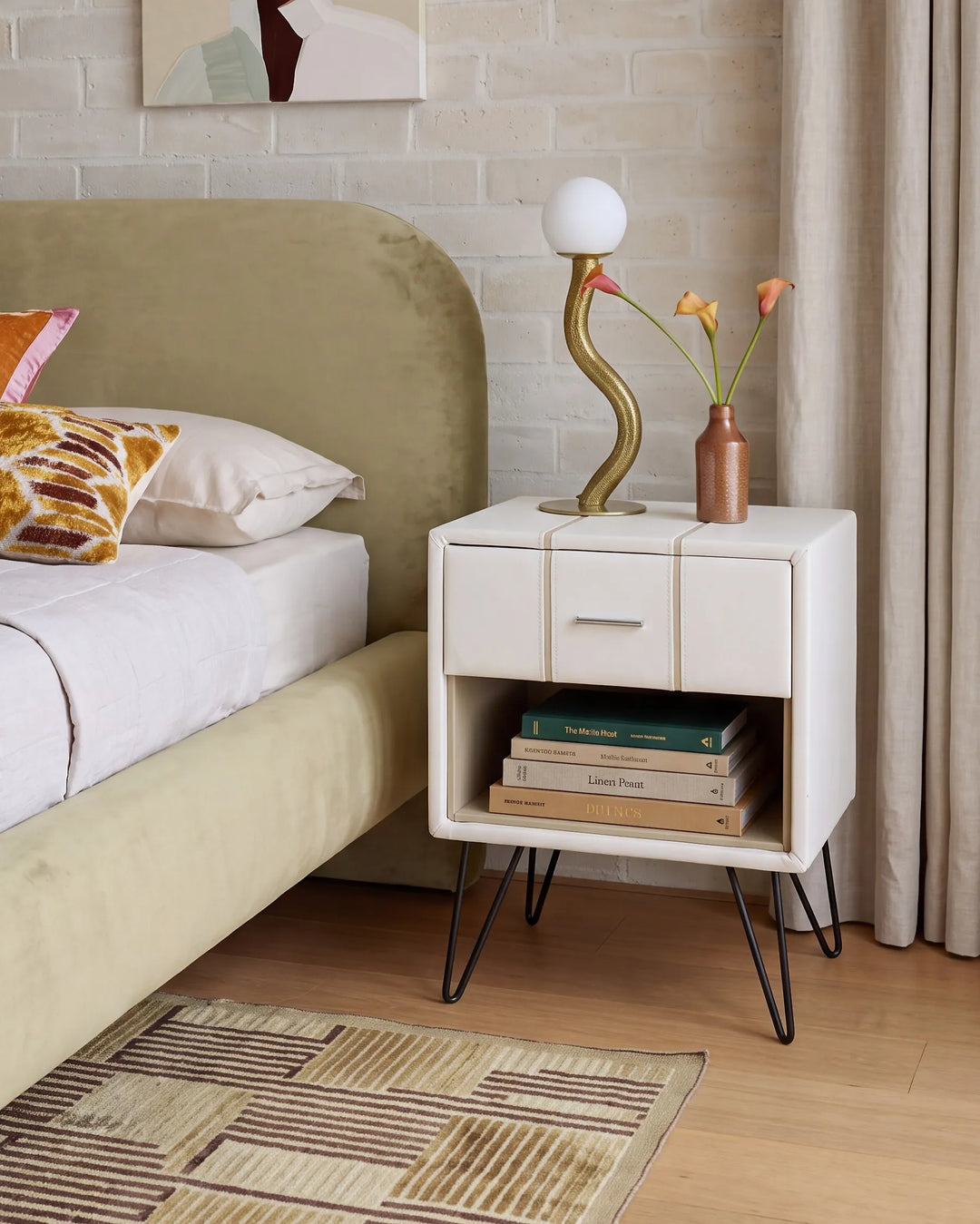 A modern white nightstand with black hairpin legs in a bedroom setting, styled with a curved gold lamp, a small vase with orange flowers, and a stack of books.