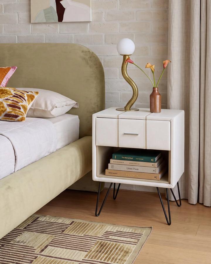 A modern white nightstand with black hairpin legs in a bedroom setting, styled with a curved gold lamp, a small vase with orange flowers, and a stack of books.