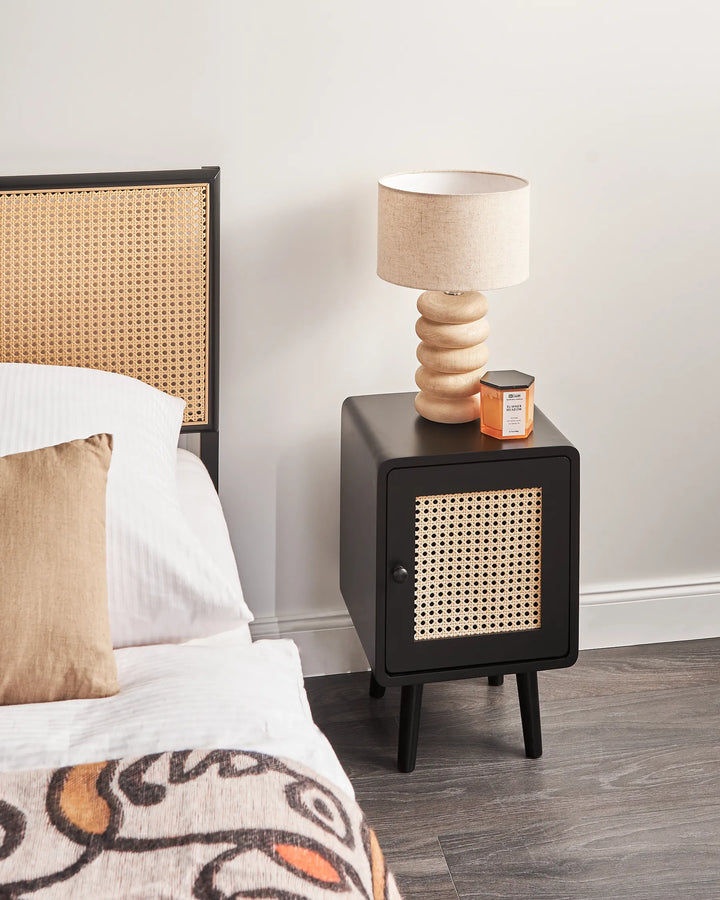 A modern black nightstand with a woven cane front panel, styled in a bedroom next to a bed with a matching cane headboard. The nightstand holds a beige fabric lampshade table lamp and a small orange candle jar.