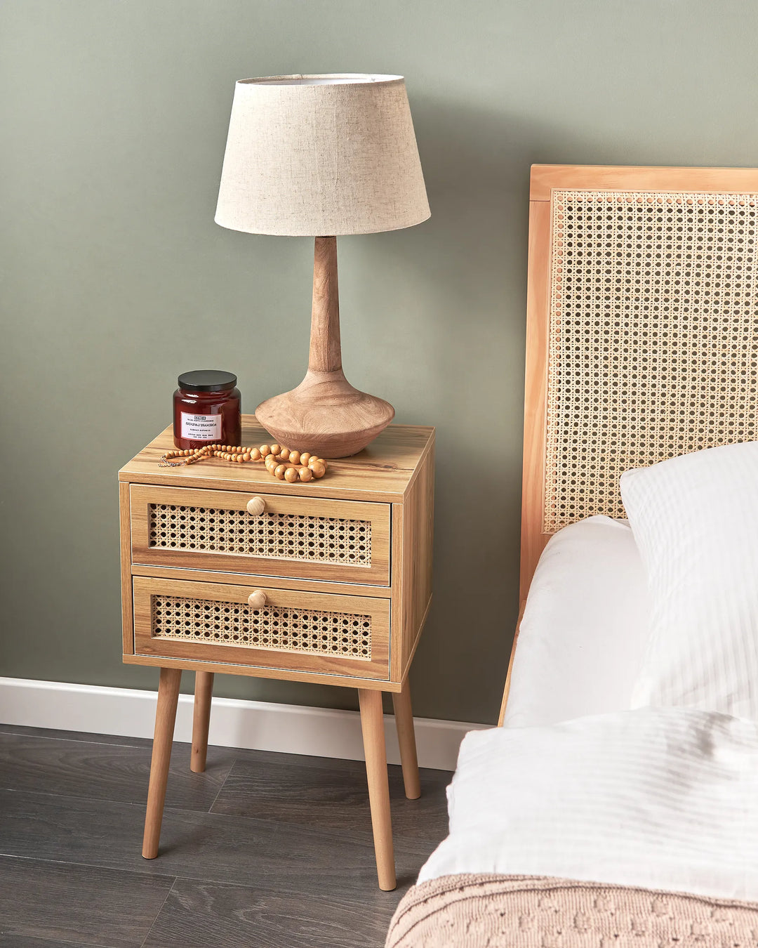 A light natural wood bedside table with two drawers featuring woven cane fronts, styled with a table lamp and a small jar in a bedroom next to a bed with a cane headboard.