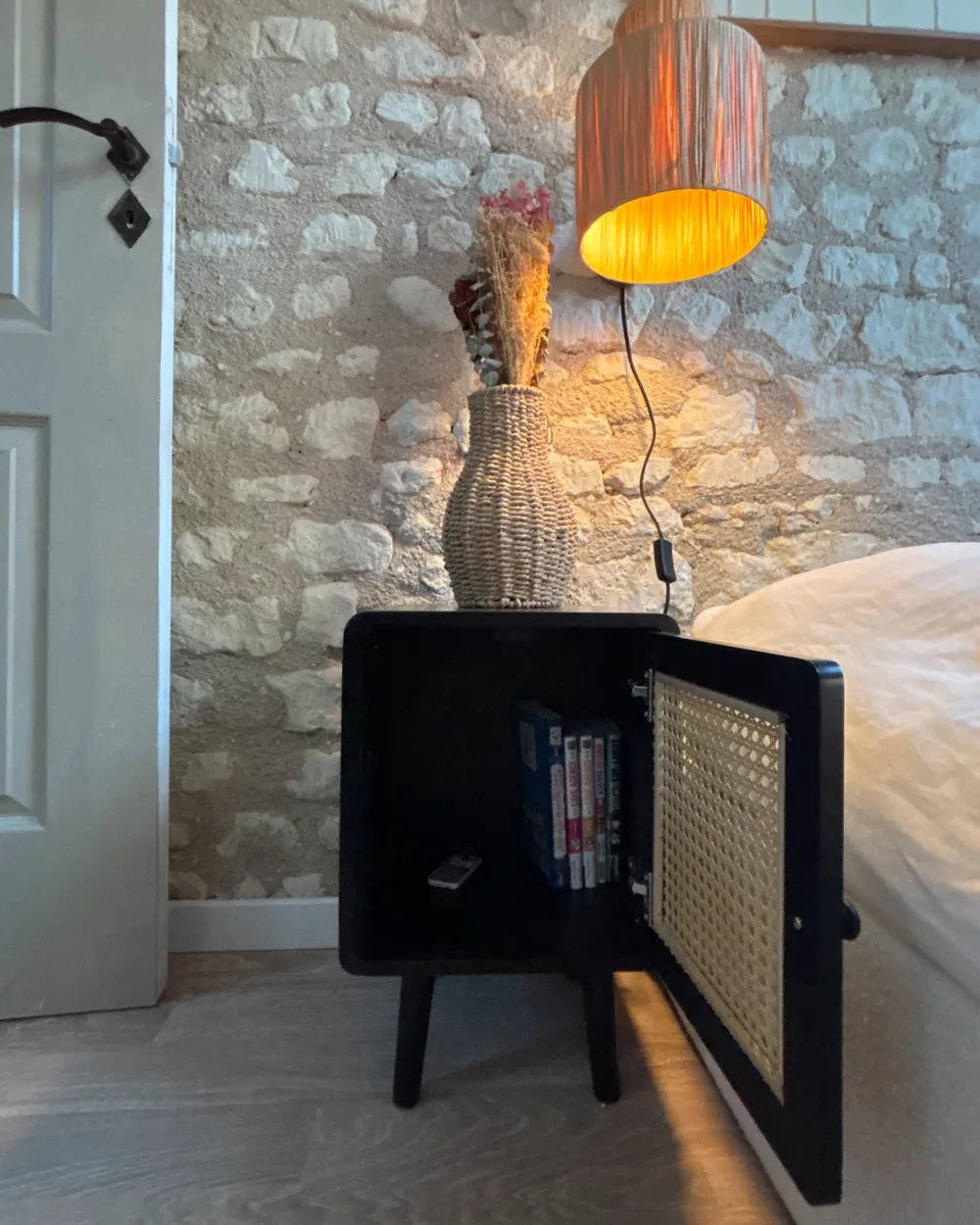 A black bedside cabinet with an open cane-look door, styled in a bedroom next to a bed with white bedding, a stone wall background, and a warm glowing lamp.