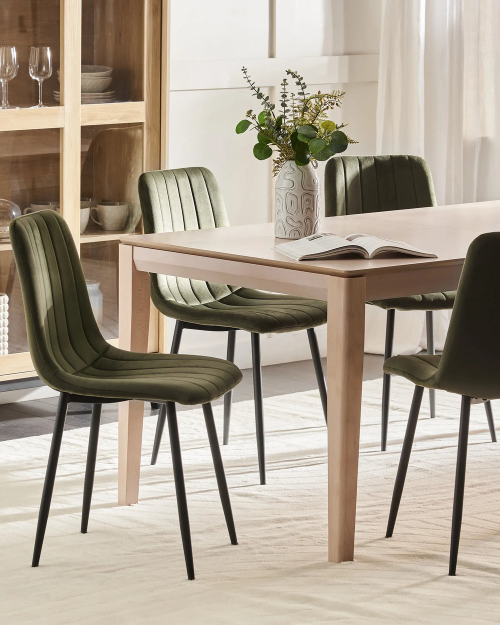 A modern dining room scene featuring a light wood rectangular table surrounded by four olive green upholstered dining chairs with black metal legs. A patterned ceramic vase with green foliage and an open book sit on the table.
