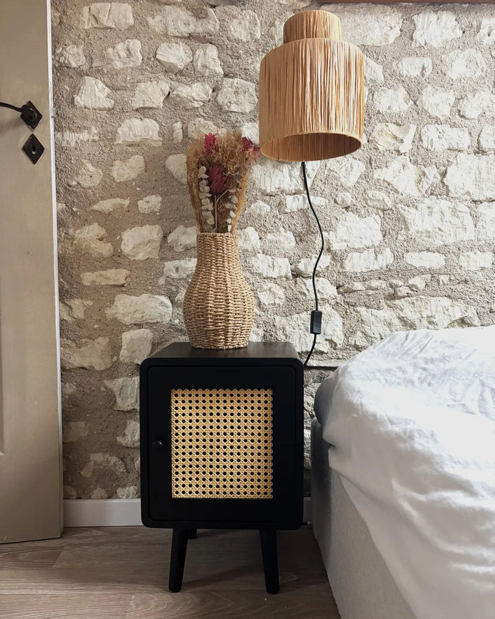 A black bedside cabinet with a woven cane front panel, styled next to a bed with white bedding and a woven lampshade lamp, set against a rustic stone wall.