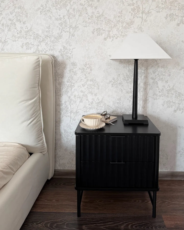 A modern black nightstand with a ribbed front and tapered legs, styled in a bedroom next to a cream-colored bed. On top of the nightstand sits a white table lamp with a conical shade, a beige ceramic cup and saucer, and a pair of eyeglasses, all set against a light gray floral wallpaper background.