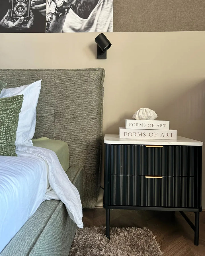 A styled bedroom corner featuring a black ribbed nightstand with a white marble top and gold drawer pulls, next to a bed with a textured green headboard. The nightstand has a stack of books reading 'FORMS OF ART' on top.