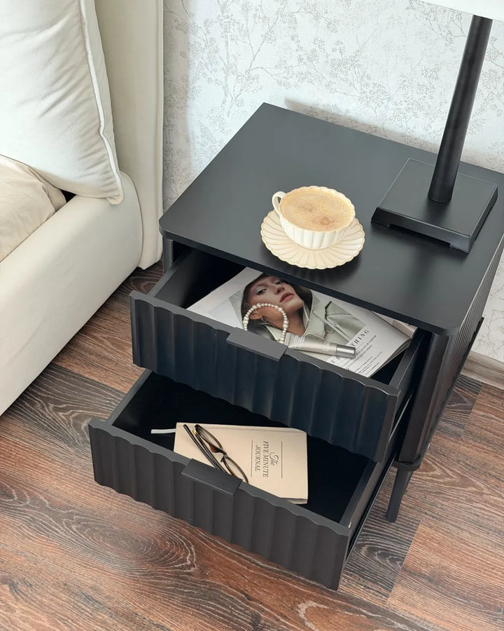 A modern black nightstand with two ribbed drawers, shown in a bedroom setting with one drawer open to reveal magazines and glasses. A coffee cup sits on top next to a lamp, with a white sofa and wood flooring visible.
