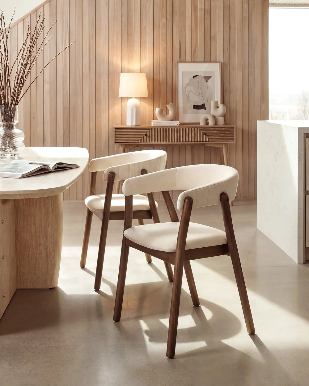 Two modern dining chairs with dark wood frames and cream upholstered seats, placed in a warm, sunlit room with wood-paneled walls and a light stone floor.