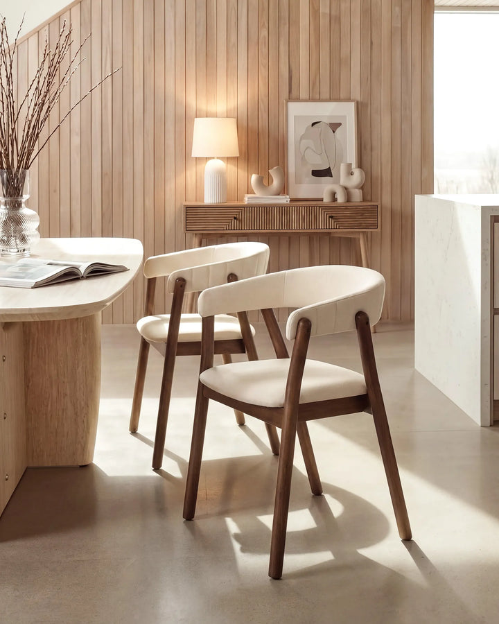 Two modern dining chairs with dark wood frames and cream upholstered seats, placed in a warm, sunlit room with wood-paneled walls and a light stone floor.